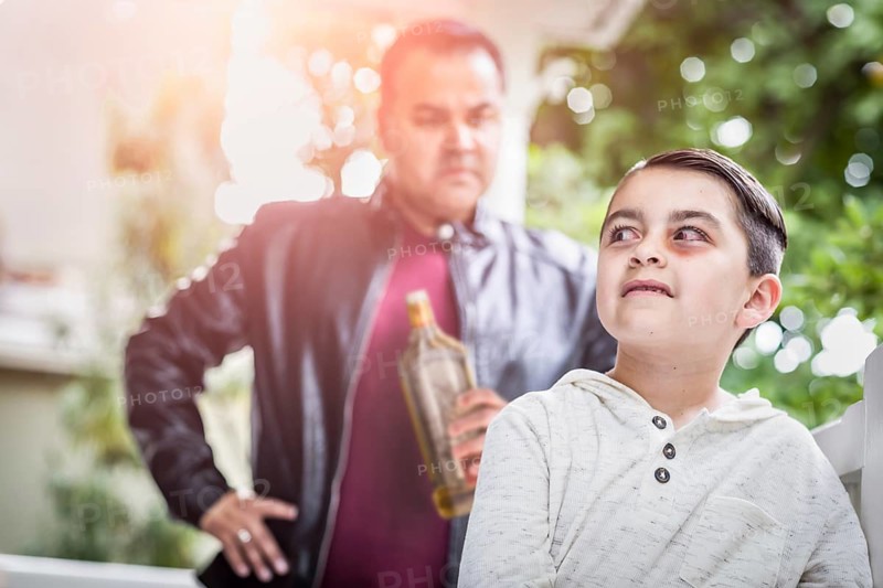 Afraid and bruised mixed-race boy in front of angry man holding bottle of alcohol