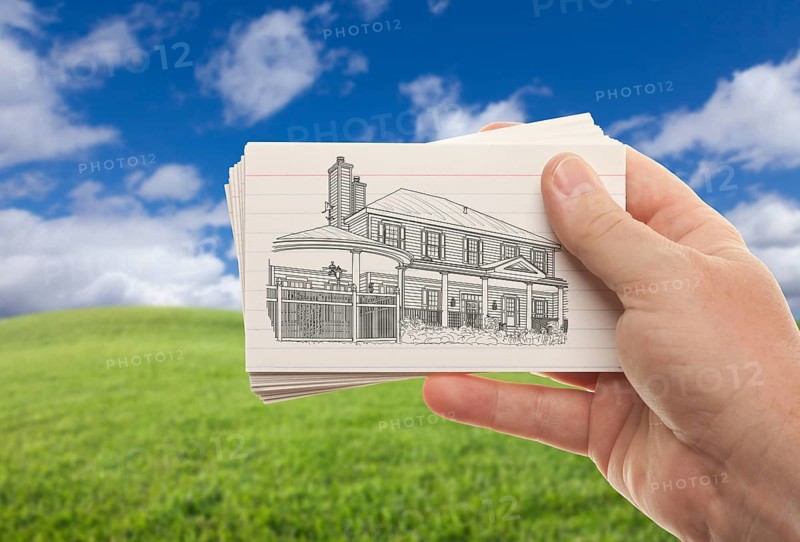 Male hand holding stack of paper with house drawing over empty grass field and sky