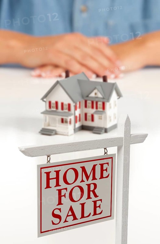 Womans folded hands behind model house and home for sale real estate sign in front on white surface