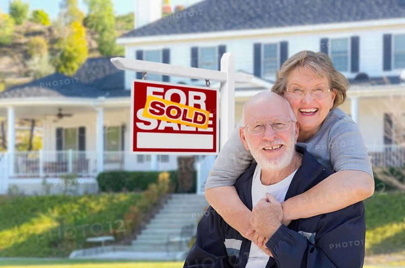 Senior adult couple in front of sold home for sale real estate sign and beautiful house