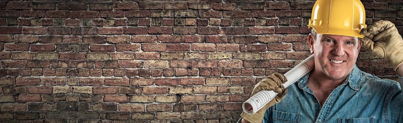 Male contractor in hard hat holding construction plans in front of old brick wall banner with copy space
