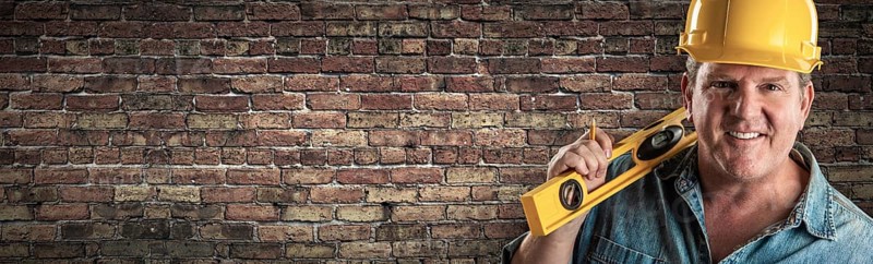 Male contractor in hard hat holding level in front of old brick wall banner with copy space