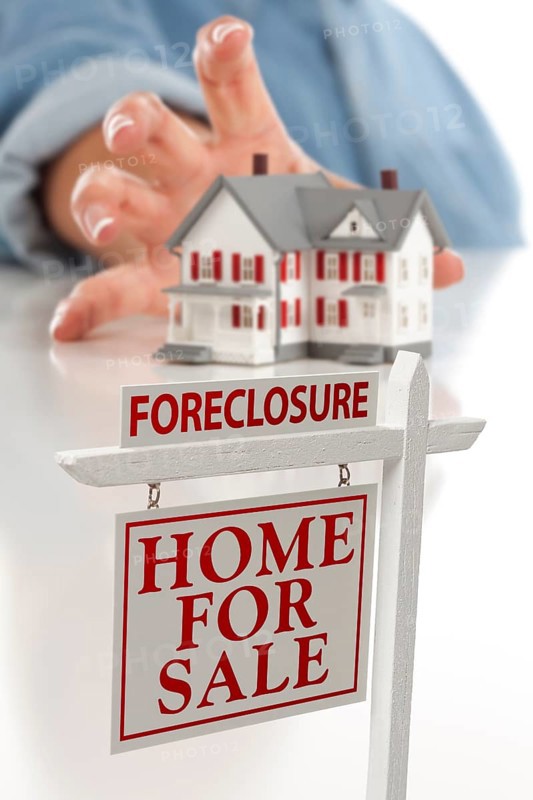 Foreclosure real estate sign in front of womans hand reaching for model house on a white surface