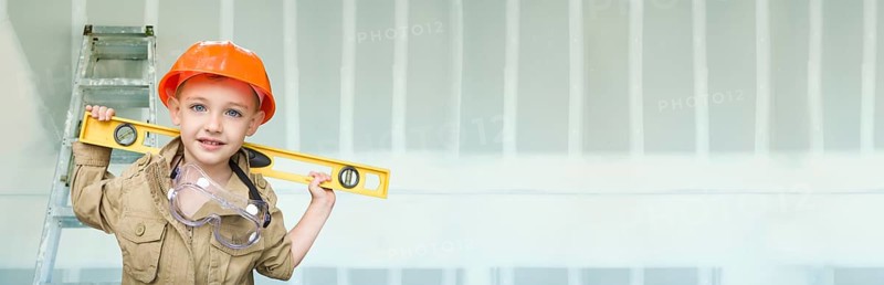 Cute young boy dressed as contractor holding level against dywall banner background with ladder