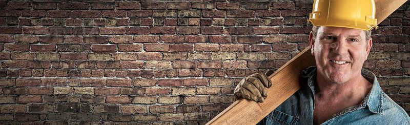 Male contractor in hard hat holding wood plank in front of old brick wall banner with copy space