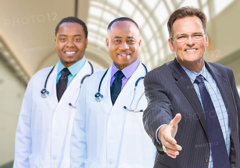 Two mixed-race doctors behind businessman reaching for a hand shake inside hospital