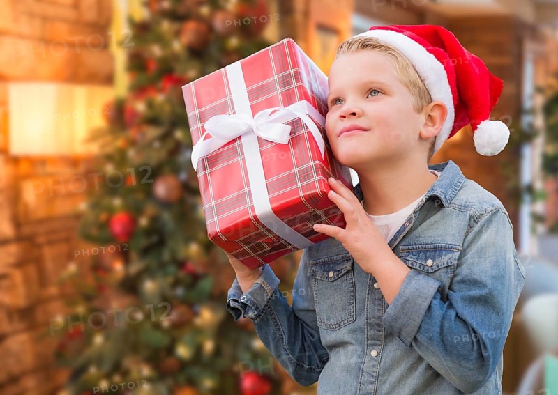 Curious young boy wearing santa hat holding christmas gift in front of decorated tree