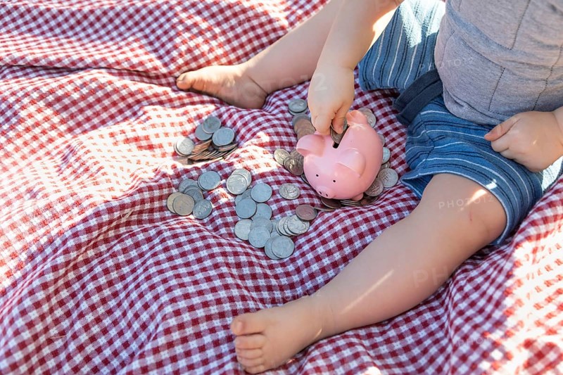 Baby boy sitting on picnic blanket putting coins in piggy bank