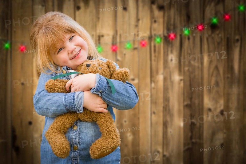 Cute little girl holding her teddy bear in front of wooded background with christmas lights