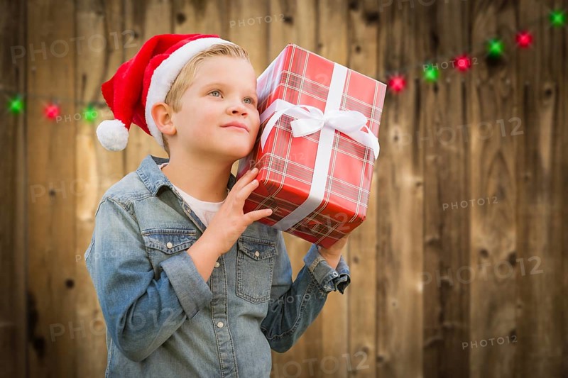 Curious young boy wearing santa hat holding christmas gift on A wood fence background