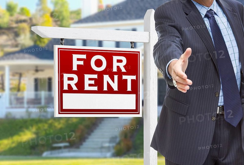 Male agent reaching for hand shake in front of for rent sign and house