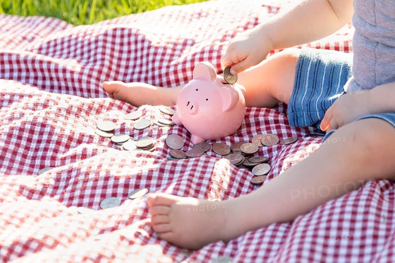 Baby boy sitting on picnic blanket putting coins in piggy bank