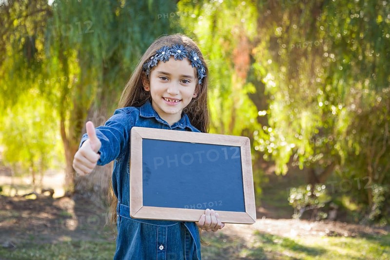 Cute young mixed-race girl with thumbs up holding blank blackboard outdoors