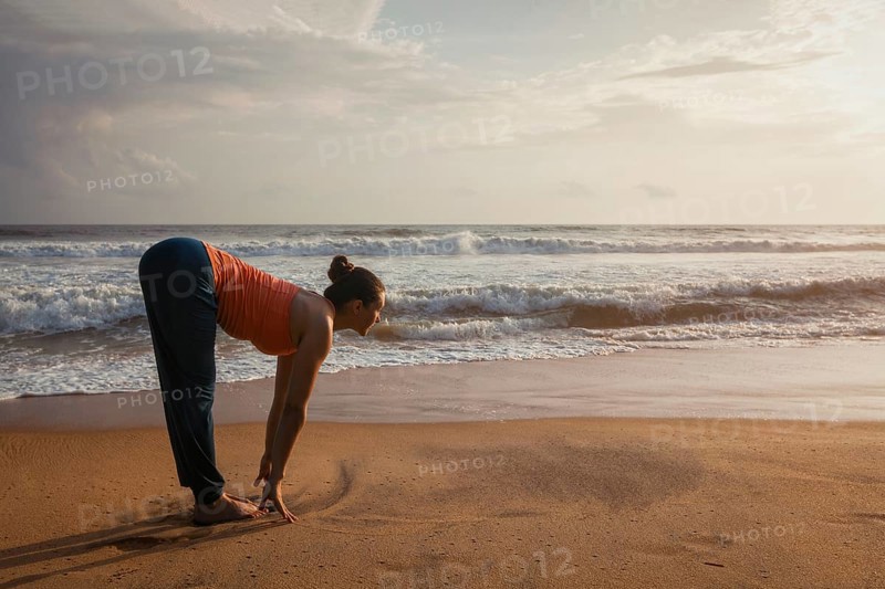 Young sporty fit woman doing yoga Sun salutation Surya Namaskar pose Uttanasana on tropical beach on sunset. With lens flare