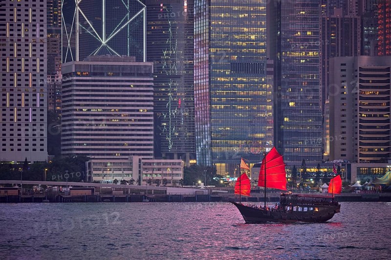 Tourist junk boat ferry with red sails and Hong Kong skyline cityscape downtown skyscrapers over Victoria Harbour in the evening. Hong Kong