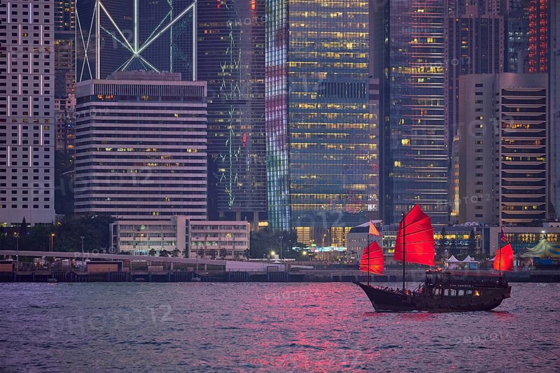 Tourist junk boat ferry with red sails and Hong Kong skyline cityscape downtown skyscrapers over Victoria Harbour in the evening. Hong Kong