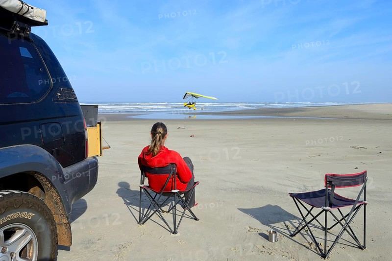 Woman sitting on camping chair next to all-terrain vehicle on beach watching a light aircraft taking off