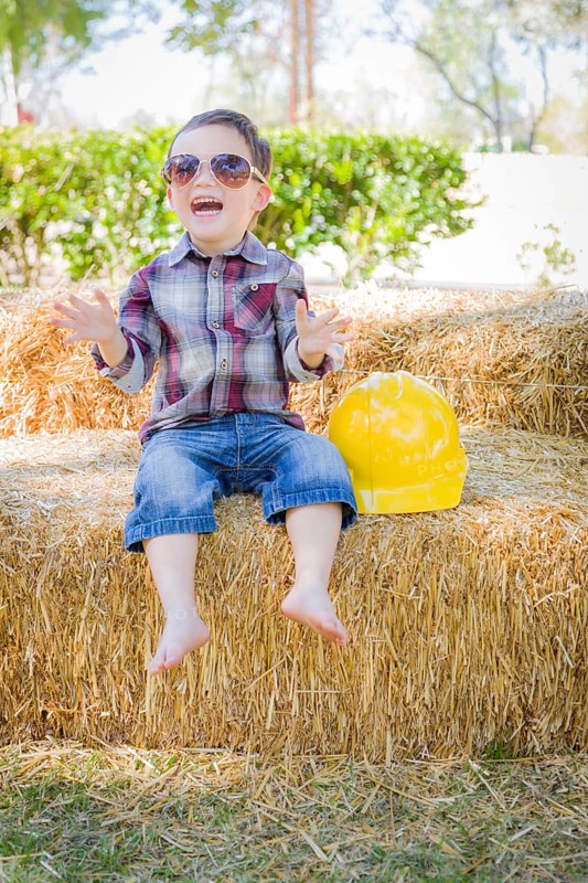 Cute young mixed-race boy laughing with sunglasses and hard hat outside sitting on hay bale