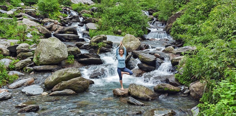 Woman practices balance yoga asana Vrikshasana tree pose at waterfall outdoors. Himachal Pradesh