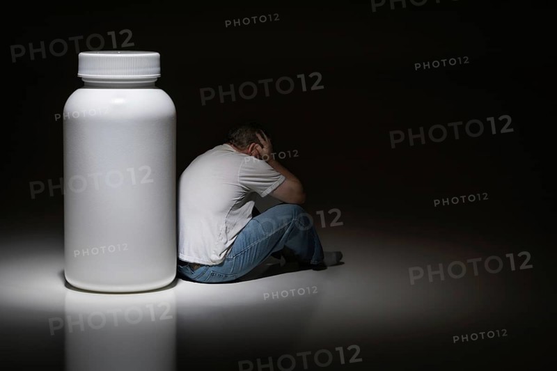 Man holding head sitting next to blank medication bottle