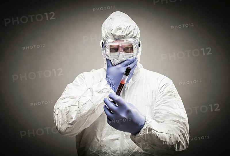 Man wearing HAZMAT protective clothing holding test tube filled with blood against A gray background