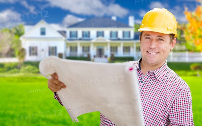 Smiling contractor holding blueprints in front of beautiful custom home