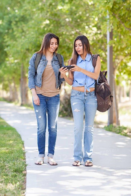 Two beautiful young ethnic twin sisters with backpacks using A smartphone outside