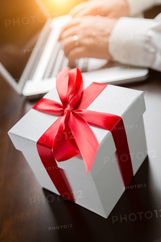 White gift box with red ribbon and bow near man typing on laptop computer