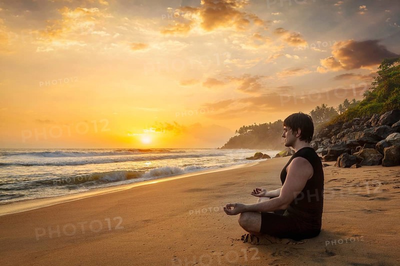 Young sporty fit man doing yoga meditating in padmasana lotus pose on tropical beach on sunset