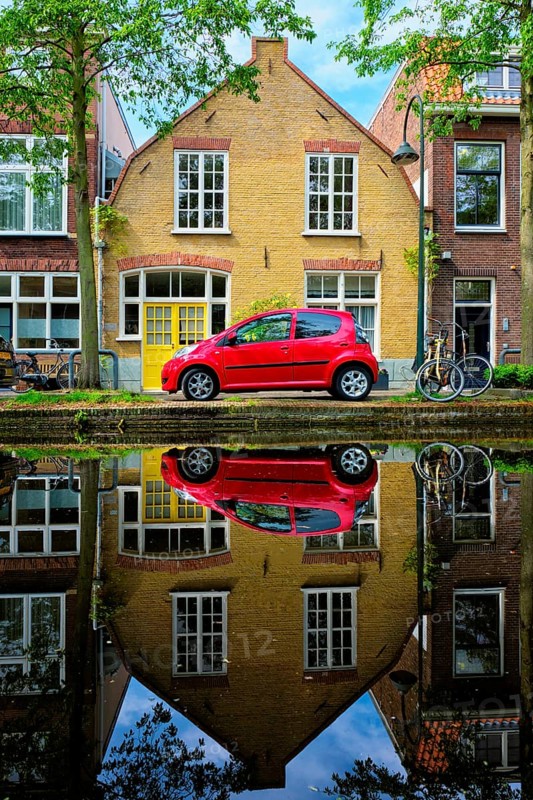 Red car on canal embankment in street of Delft with reflection and bicycle