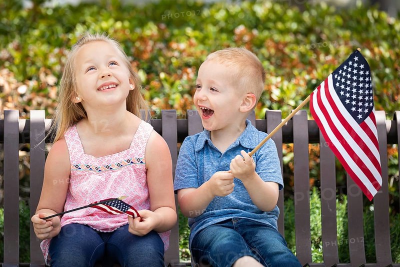 Young sister and brother comparing each others american flag size on the bench at the park