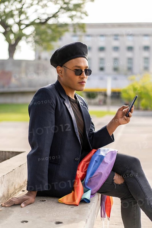 Latino gay male with makeup on wearing trendy hat holding a cellphone and lgbt flag in a park