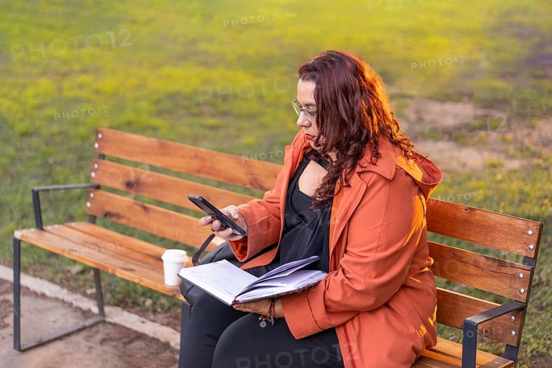 Woman sitting on a bench in a park checking her agenda and cell phone while having a coffee