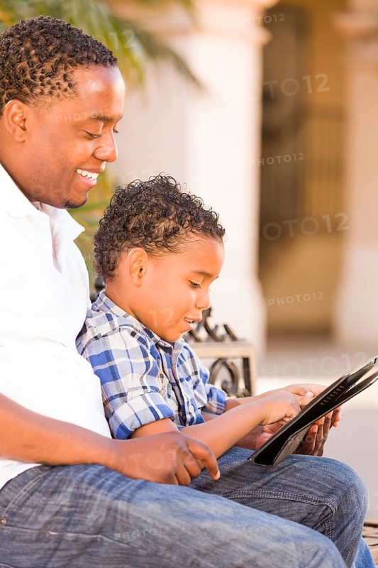 African american father and mixed-race son using computer tablet on bench in park