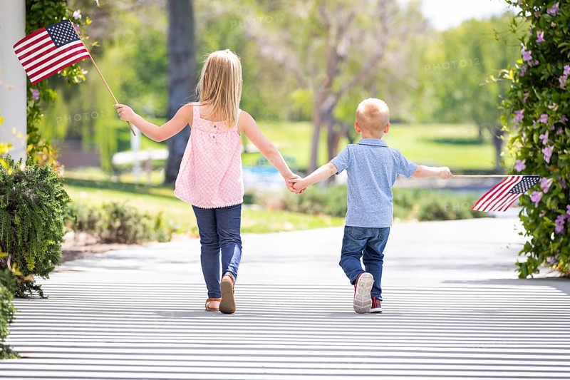 Young sister and brother holding hands and waving american flags at the park
