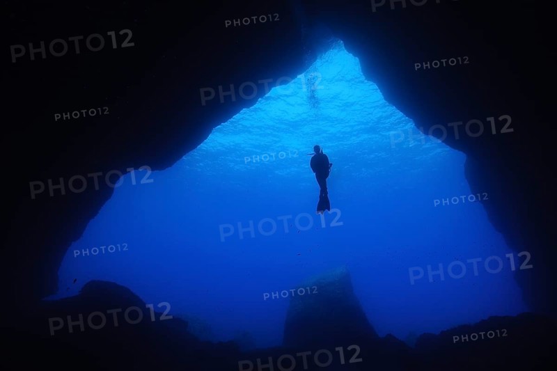 Diver floating in open water in front of large cave entrance of underwater cave