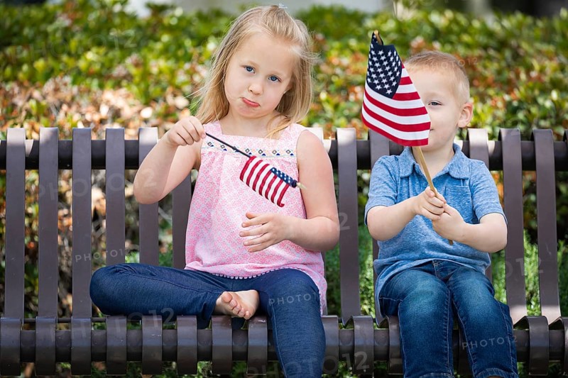 Young sister and brother comparing each others american flag size on the bench at the park