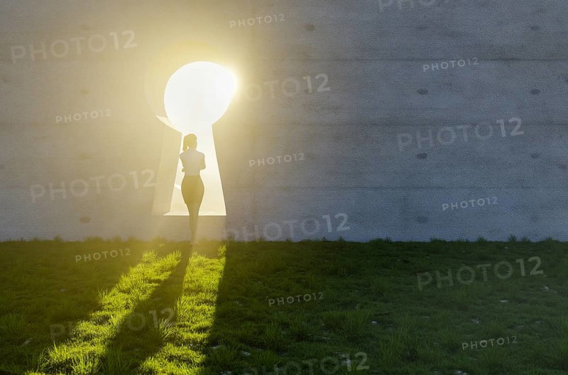 Woman standing at bright sun light shining through keyhole in concrete wall in grass and rocks field