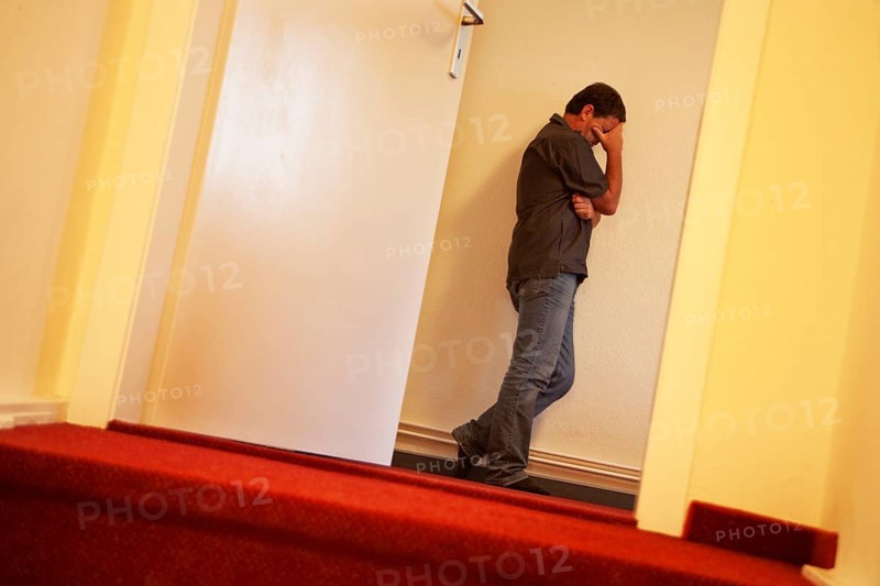 Verzweifelter Mann leht an einer Wand im Stiegenaufgang hiner einer Tuere