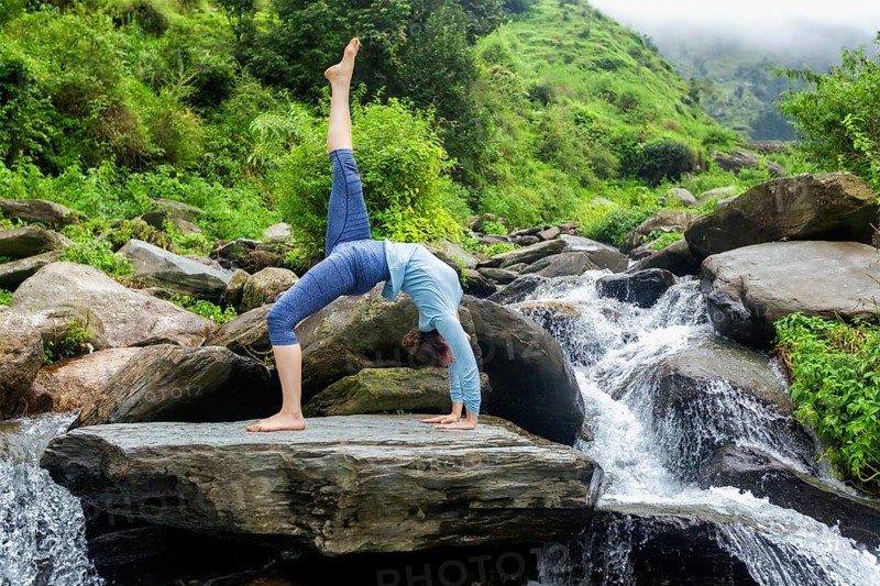 Woman doing yoga asana eka pada urdva dhanurasana Upward Bow Pose outdoors at waterfall in Himalayas
