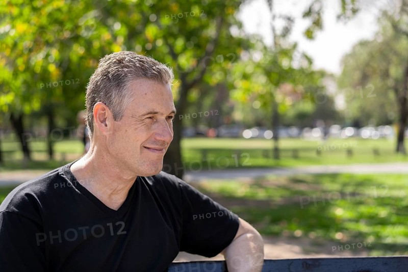Close-up portrait of a mature man with gray hair wearing a black t-shirt sitting on a park bench looking to the side