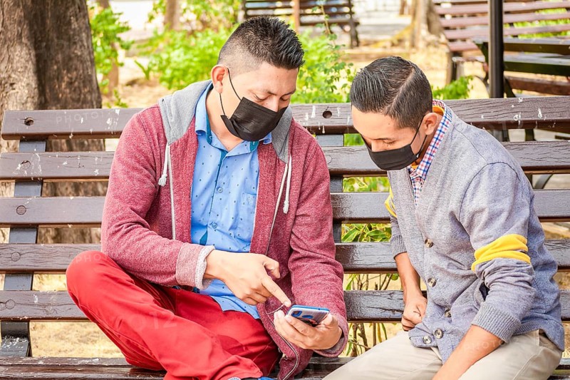 Two men wearing masks checking their cell phones on a bench