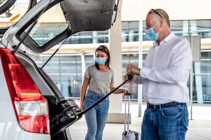 A taxi or Uber driver helping a passenger in a protective mask with her luggage at the airport
