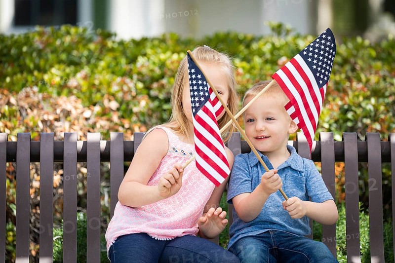 Young sister and brother waving american flags on the bench at the park