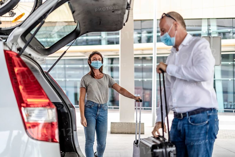 A taxi or Uber driver helping a passenger in a protective mask with her luggage at the airport