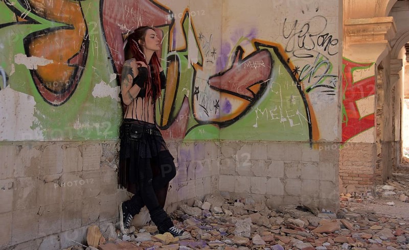 Young woman with tatoos and dreadlocks stands against wall with graffiti of an abandoned house