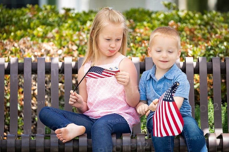 Young sister and brother comparing each others american flag size on the bench at the park