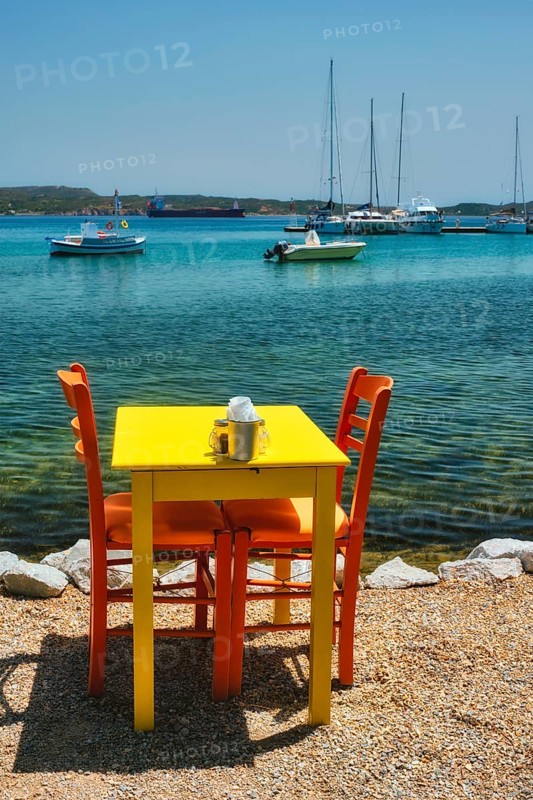 Yellow cafe restaurant table of street cafe with chairs on beach in Adamantas town on Milos island with Aegean sea with boats and yachts in background. Milos island