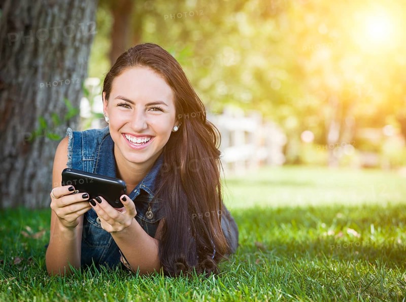 Attractive happy mixed-race young female laying in the grass texting on her cell phone outside
