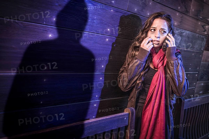 Scared young adult woman uses cell phone as mysterious male shadow figure lurks nearby